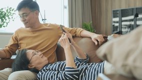 Happy senior old asian lover couple holding smartphone looking at cellphone screen laughing casual relaxing sit on sofa together, smiling elder mature grandparents family embracing lifestyle