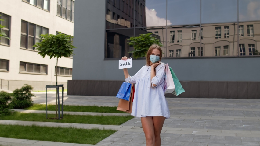Young stylish model girl in protective mask with shopping bags showing Sale word inscription text, looking satisfied with low prices purchases during coronavirus, covid-19 pandemic. 6k downscale | Shutterstock HD Video #1059328676