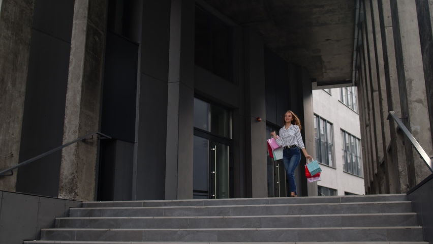 Attractive girl in shirt and jeans with shopping bags, spining, dancing, looking satisfied with purchase, enjoying price discounts on Black Friday sale holiday. Central mall stairs. 6k downscale
