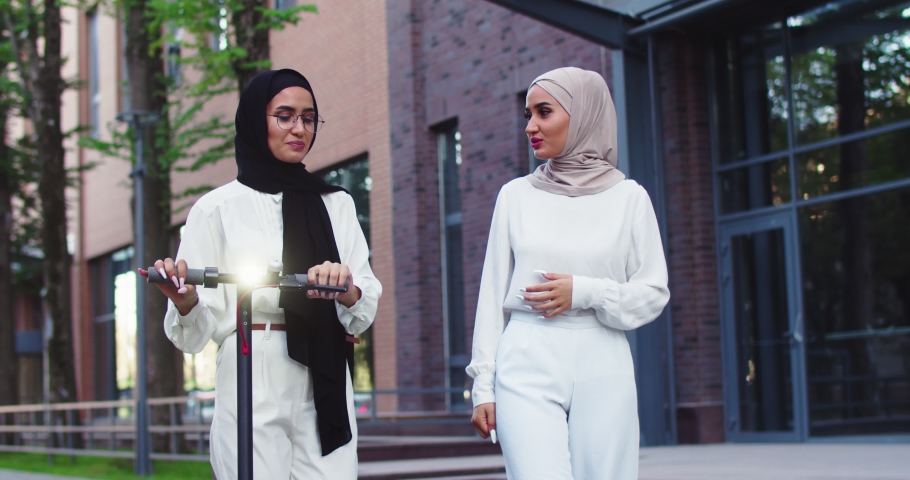 Middle plan shot of two muslim beautiful women in traditional headscarves talking, walking on street with personal e-scooter near beautiful urban building. Pretty Arabian females in hijabs at city.
