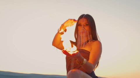 Beautiful young woman holding fire in her hands. An adult woman with a creative oriental make-up, her face is hidden behind a golden mask. Fashion model posing against the sky. video 4k