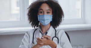 Female African American woman nurse in medical mask sitting in clinic, doctor talking on camera online video conference chat, active moving gesticulate with hands, covid19 virus concept, webcam view