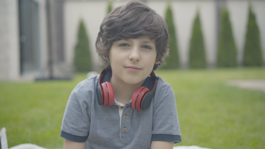 Portrait of cute brunette kid showing thumb up and laughing at camera. Cheerful Caucasian boy posing outdoors. Relaxed happy child enjoying weekends at home.
