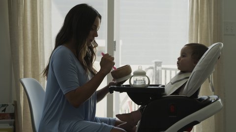 Mother feeding baby daughter in high chair near window / Bluffdale, Utah, United States
