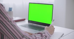 young woman is learning, writing in copybook and viewing green screen on laptop for chroma key technology