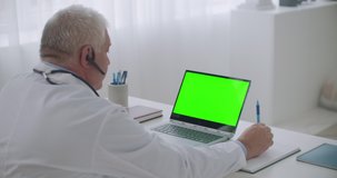 male doctor is consulting online from his office, view on laptop with green display for chroma key technology, looking at screen