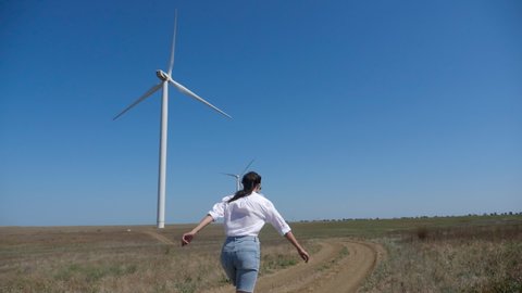 young woman in a shirt and denim shorts walks across the field with a wind turbine