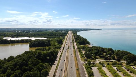Aerial view of cars driving on a highway next to a beautiful emerald lake. Top view of a cars driving on a highway through green natural park between Lake Ontario and Sixteen Mile Pond in Canada.
