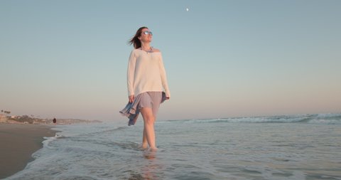 Cinematic slow motion woman enjoying nature at sunset with blue and rose sky and Moon in the twilight, 4K. Attractive female in wavy dress walking barefoot by the beach with ocean breeze blowing hair 