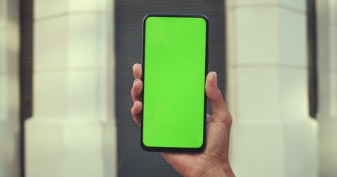 Close up view of female hand holding mobilephone and pressing on mockup blank screen. Concept of greenscreen and chroma key. Closed building at background