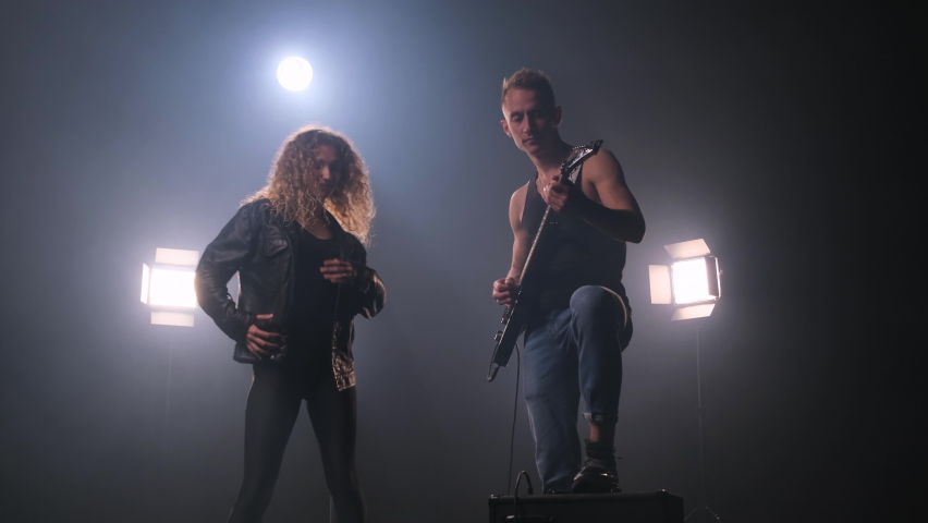 Two rock stars on stage. The guy in the black T-shirt plays the electric guitar. Woman with lush curly hair in leather jacket dances around him and enjoys music. fog around