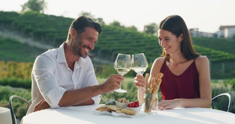 Authentic shot of happy couple in love is enjoying romantic dinner together and cheering with white wine glasses to celebrate their anniversary and timeless love on scenic vineyards background.