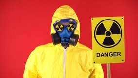 A man in a protective chemical suit shakes his head negatively with a poster of Radiation. Isolate, studio shooting. Radiation hazard concept on red background.