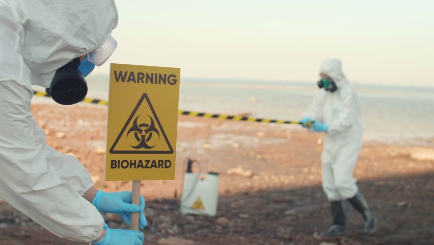 Panning of two scientists wearing chemical suits cordoning off territory where environmental collapse has happened. Scientist installing warning sign and rolling out warning tape Royalty-Free Stock Footage #1059377123