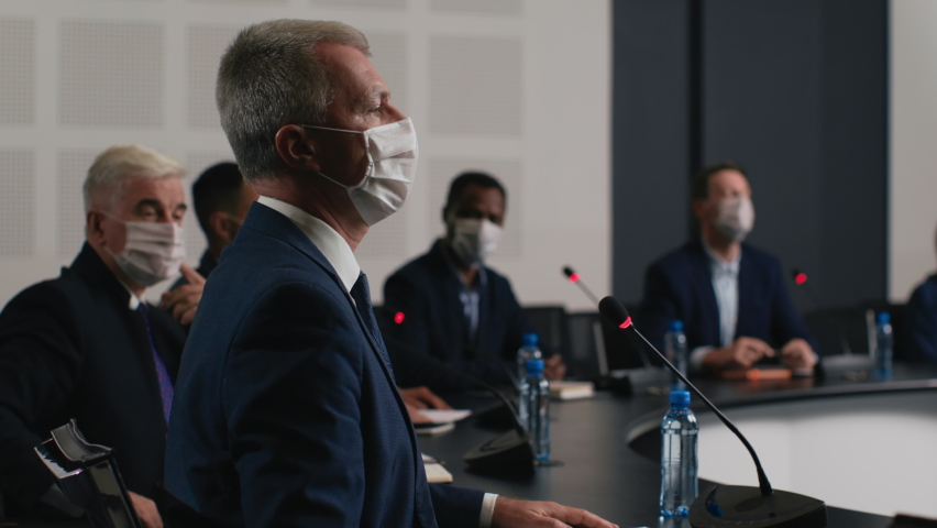 Political speaker talks at meeting room of business center. Man in suit and mask explains coronavirus epidemic. 2019-ncov conference indoors of convention hall. Expert group works at official event Royalty-Free Stock Footage #1059381560
