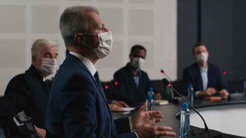 Political speaker talks at meeting room of business center. Man in suit and mask explains coronavirus epidemic. 2019-ncov conference indoors of convention hall. Expert group works at official event