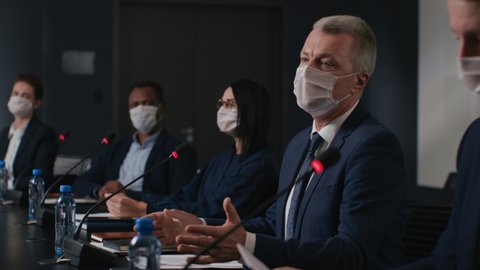 Speech of political speaker at meeting room of business forum. Man in suit and mask discusses covid-19 pandemic. 2019-ncov conference indoors of convention hall. Expert group works at official event