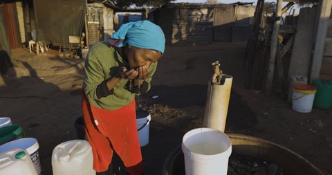 Face of poverty.  Poor Black African woman with no access to running water, drinks water while filling up buckets at a central tap to take to her house in an informal settlement (squatter camp) 