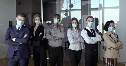 Motivated multidimensional business team stands together in conference room, confident colleagues fold arms over chests, office workers wear protective medical masks preventing virus, safety concept