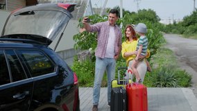 happy joyful male and female parents with their beloved son in their arms talking via video link with friends, near the suitcases for family trip by car during summer vacation