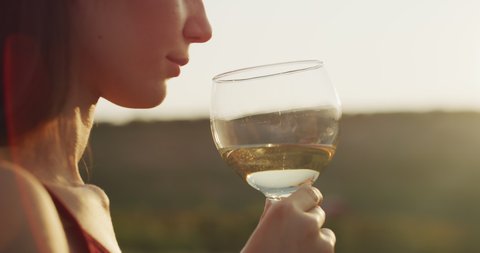 Authentic shot of happy young elegantly dressed woman is tasting a flavor and drinking a white wine poured in transparent glass on scenic vineyards background at sunset.