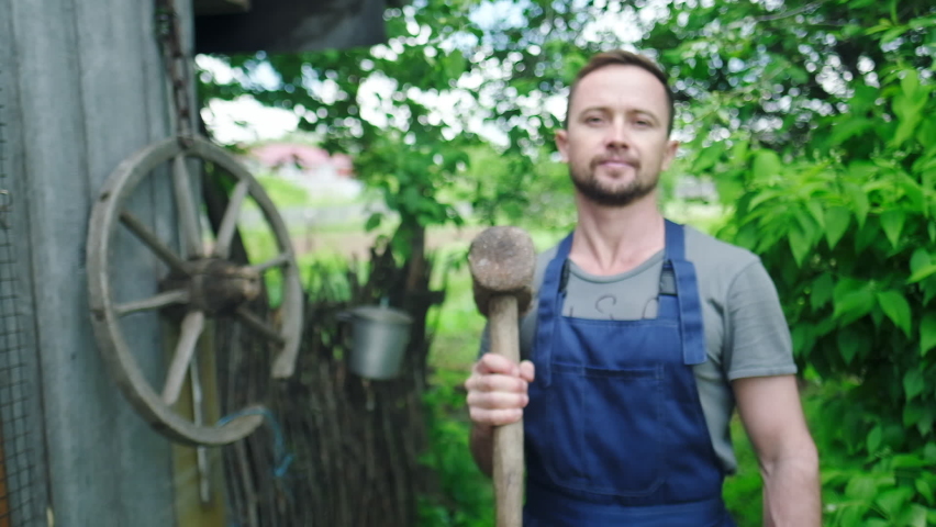 Playful worker in the garden. Young adult man plays with heavy hammer in the garden and looks at the camera
