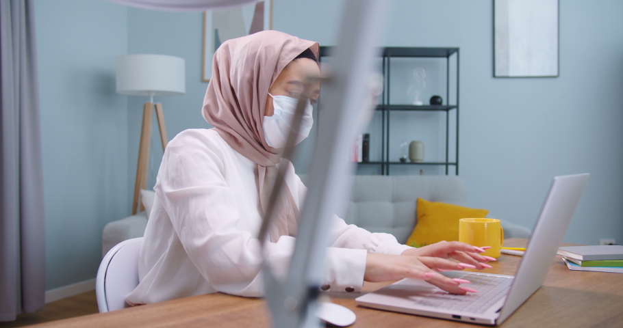 Middle plan of sick young businesswoman in hijab wearing face mask typing on computer, searching in Internet at home office. Muslim female working distantly self-isolating having coronavirus symptom. Royalty-Free Stock Footage #1059410747