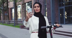 Young Arabic beautiful women in traditional headscarves, influencer shooting video blog, making selfie with electric scooter, meeting a friend on the street. Pretty Arabian females in hijabs at city.