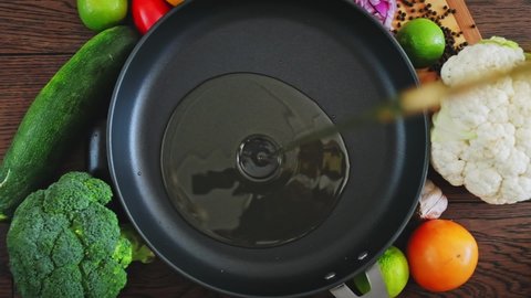 Cook pours sunflower oil into a frying pan, top view. Kitchen pan stands on brown boards, vegetables around it, and oil is poured into it for frying.  Slow motion  of  oil pouring into a frying pan. 
