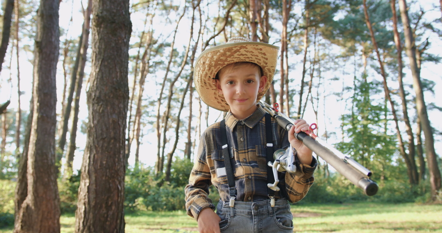 Likable carefree joyful 10-aged boy wearing hat posing on camera with fishing tackles in the middle of green afforestation during going fishing