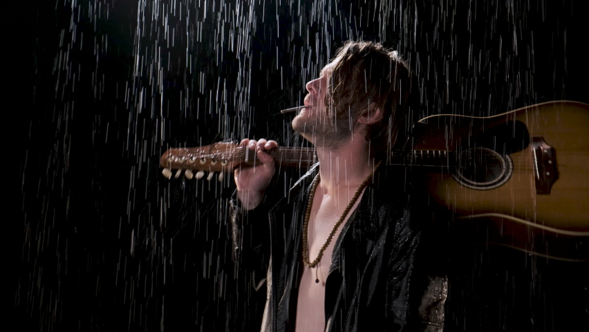 Man with long blond hair with guitar on his shoulder and cigarette in his mouth is wearing black leather biker jacket in the rain. The concept of sad singing under his girlfriend window.