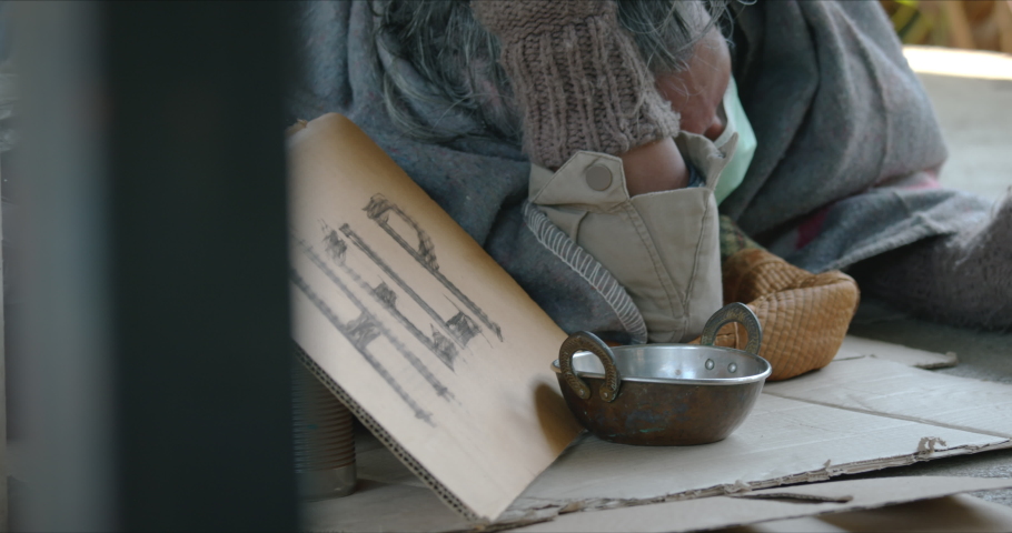Homeless senior man with dirty clothes and old blanket is lying down on the sidewalk is holding a cup to ask for money and help from kindly people.
