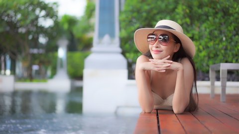 While lying prone on a wooden deck surrounding a large resort pool, a young woman in a sun hat and sunglasses leaning on her hands smiles as she looks out across the hotel property.