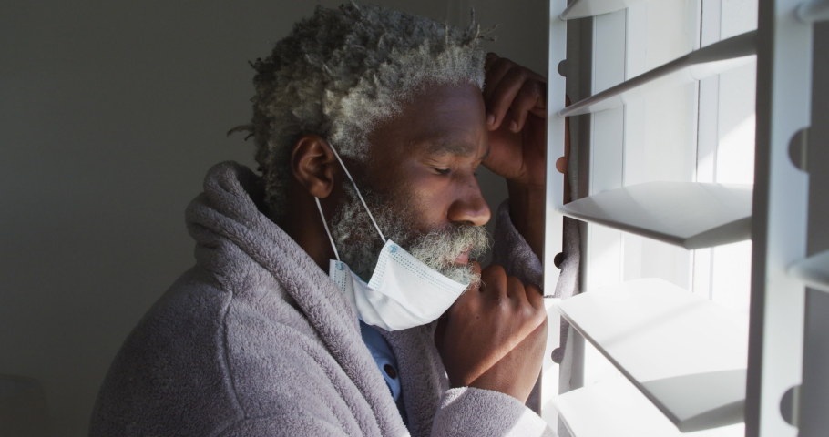 African American senior man standing in a bedroom, wearing a face mask, looking through a window, social distancing and self isolation in quarantine lockdown, slow motion