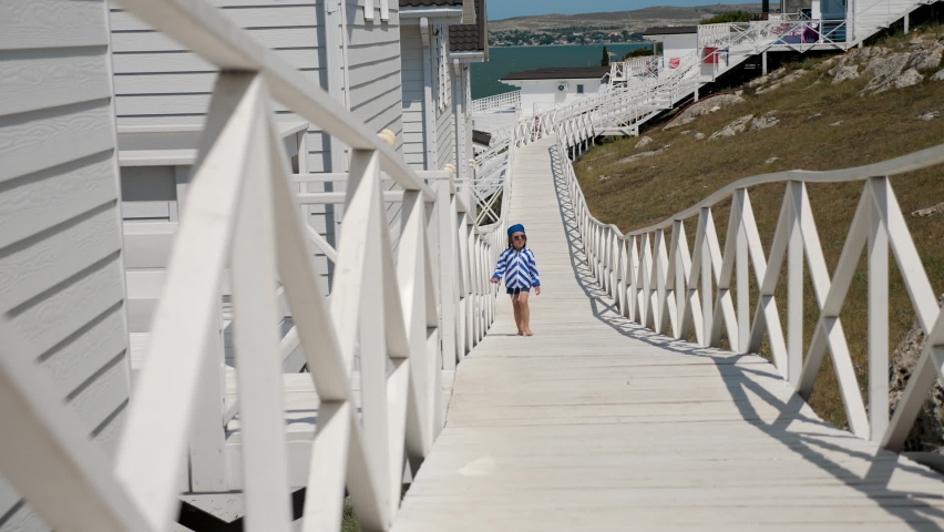 boy child in a striped blue jacket and bandana walks on a wooden platform among white houses by the sea