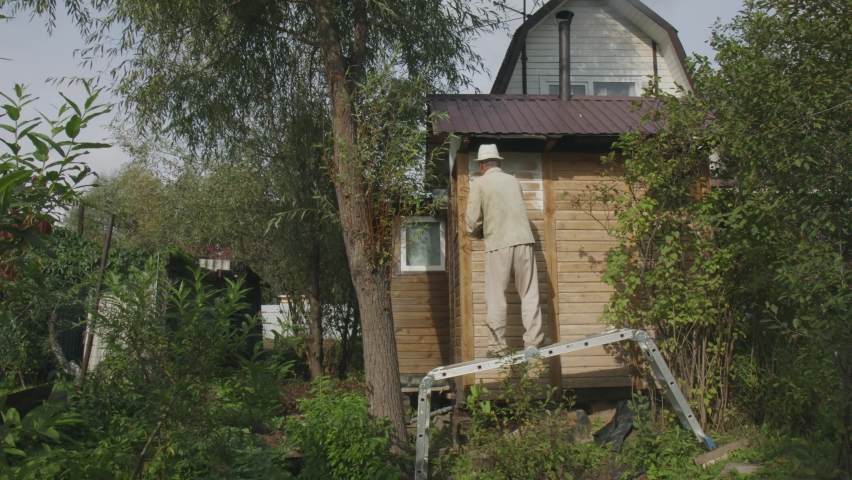 An adult man standing on the stairs repairs house in the open air, paints the exterior walls with white paint: lifestyle, home renovation and renovation concept. Painting. Rear view, slow motion, 4K