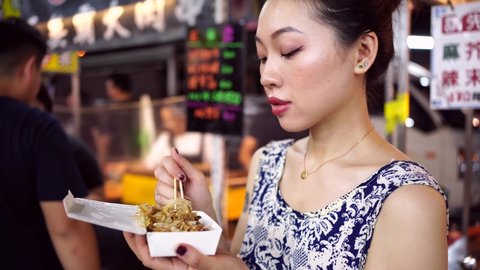 Ethnic female standing in Zhubei Night Market and blowing on hot octopus balls while enjoying Asian food with chopsticks