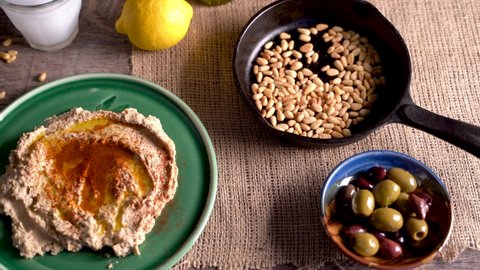 High angle slide to the left of middle eastern food spread with hummus, pine nuts, lemons, olives, get and ouzo or arak.
