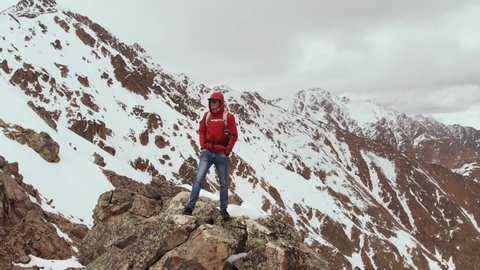 A male traveller with a backpack stands on top of a cliff against the backdrop of snow-capped mountain peaks and low clouds. Aerial view. Flying the camera around a man in a storon cliff