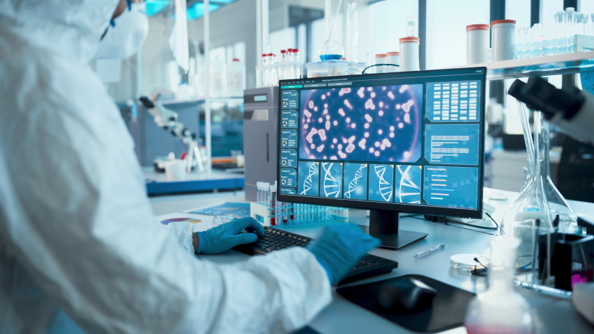 Medical Laboratory: Microbiology Scientist Wearing Sterile Coverall, Face Shield and Mask Works on Computer Developing Vaccine, Drugs and Antibiotics. Screen Shows Concept for DNA research. Zoom out