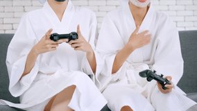 Two happy Asian girls in white bathrobes with facial mask playing video game together in living room.