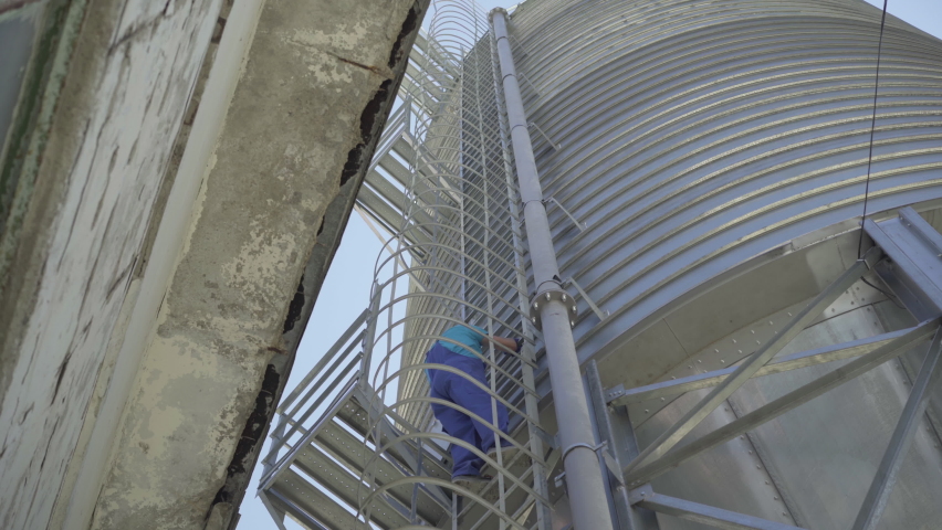 Wide shot of man in uniform and safety helmet walking on enormous cement storage silo. Huge stainless steel tower with Caucasian factory worker climbing up using ladder.