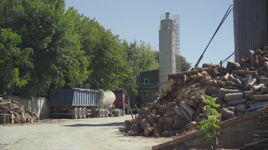 Wide shot of industrial manufacturing site with cement storage silo and felled tree logs lying outdoors in sunlight. Factory producing concrete in sunrays.