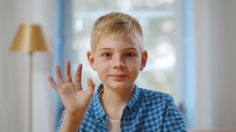 Happy cute little vlogger waving hand saying hello hi looking at camera talking to webcam. Smiling kid boy making online video call recording vlog sitting at home