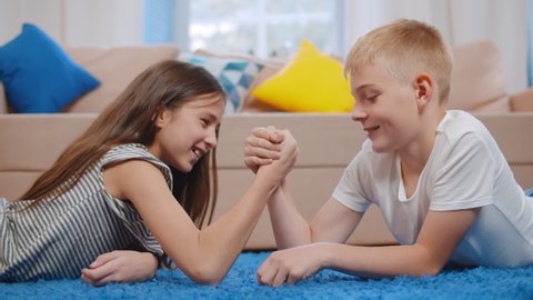 Happy boy and his sister having fun arm wrestling laying on the floor in living room. Portrait of cheerful caucasian siblings arm-wrestling relaxing on blue carpet at home.