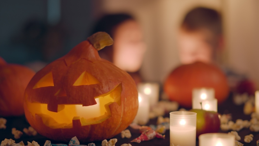 Carved Halloween pumpkin lantern on table with burning candles and sweets in decorated home with kids on blurred background. Seasonal decoration interior with pumpkins