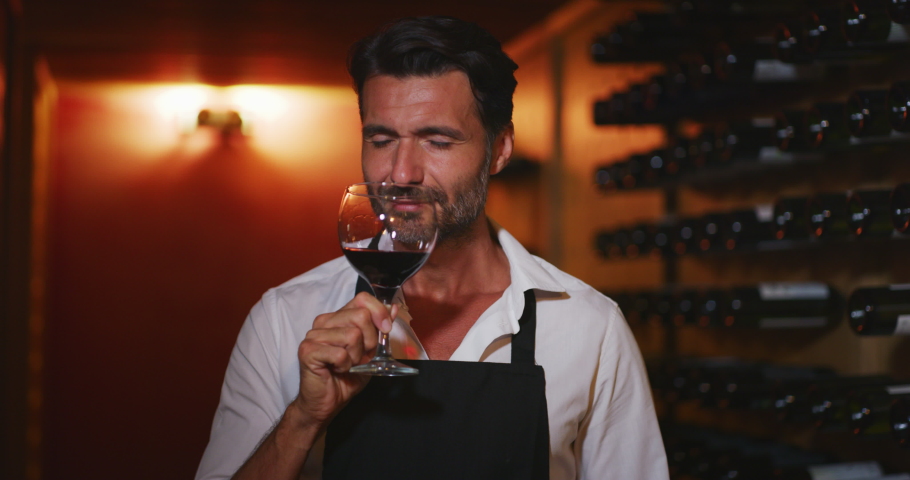 Authentic shot of happy successful male winemaker is tasting a flavor and checking red wine quality poured in transparent glass  in a wine cellar.