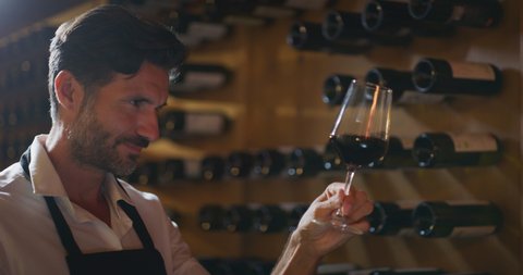 Authentic shot of happy successful male winemaker is tasting a flavor and checking red wine quality poured in transparent glass  in a wine cellar.