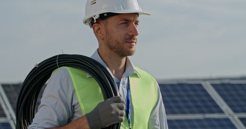 Close up view of handsome male engineer in uniform walking at solar power plant. Caucasian man in hard helmet examining object and carrying cabble. Concept of green energy