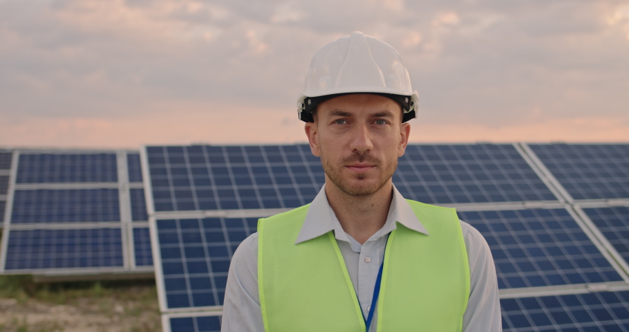 Portrait of happy male engineer in protective helmet crossing arms while looking to camera. Handsome man in uniform smiling while standing at solar power farm. Concept of green energy | Shutterstock HD Video #1059465863